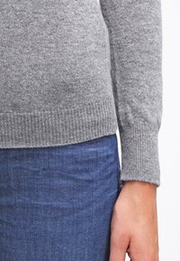 Person som bär en grå stickad tröja med ribbade muddkanter och nederkant, i kombination med blå denimjeans, med handen avslappnad vid sidan.