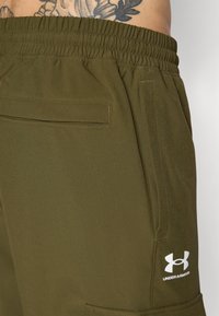 Olive green shorts with an elastic waistband. Features side pockets and a white logo on the lower left leg. Smooth, breathable fabric.