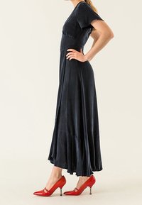 Tmavě modré maxi šaty s krátkými rukávy, přiléhavým tělem a rozšířenou sukní, doplněné červenými lodičkami s špičatou špičkou a kovovým akcentem.