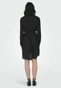 Abito nero a maniche lunghe con vita aderente, retro plissettato e orlo al ginocchio. Tessuto liscio con una sovrapposizione trasparente, abbinato a tacchi neri.