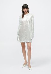 Διάφανο, μακρυμάνικο φόρεμα από υλικό με αντανακλαστικές παγιέτες σε ανοιχτό ασημί χρώμα. Ευθεία γραμμή με ελαφριά υφή. Συνδυασμένο με λευκά, μυτερά παπούτσια.