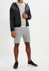 Chaqueta negra ligera con capucha, camisa gris de manga corta y pantalones cortos grises. Zapatillas negras completan el look atlético.