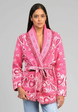 Femme portant un cardigan ceinturé à motifs roses avec des franges, par-dessus un jean bleu clair, debout sur un fond gris uni.