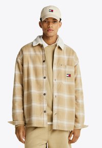 Chaqueta de camisa de cuadros beige con un suave cuello blanco, bolsillos frontales y botones negros; lleva puesta sobre un suéter beige y pantalones a juego.