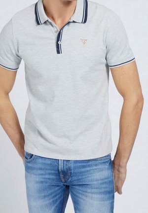 Man wearing light gray polo shirt with navy trim and small logo on chest, paired with blue jeans, standing against plain background.