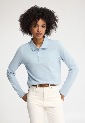 Femme aux cheveux bouclés portant un polo à manches longues bleu clair rentré dans un pantalon blanc taille haute avec une ceinture marron.