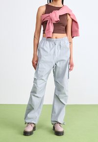 Person wearing light gray cargo pants, brown cropped tank top, pink sweatshirt tied around shoulders, and beige platform boots on green floor.