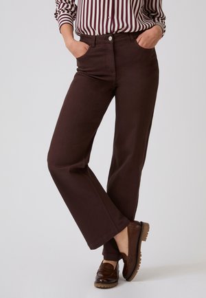 Donna che indossa pantaloni marroni a gamba dritta con le mani nelle tasche, abbinati a mocassini marrone scuro e una camicia a righe con le maniche arrotolate.