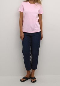 T-shirt en coton rose à manches courtes et col rond, associé à un jean bleu foncé et des tongs noires. Tenue simple et décontractée.