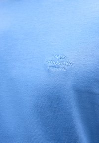 Ανοιχτό μπλε ύφασμα με μικρό λευκό κεντητό κείμενο που γράφει «MADS NØRGAARD COPENHAGEN» στο κέντρο του κάδρου.