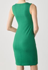Femme debout, les mains derrière le dos, portant une robe verte sans manches, ajustée et texturée, sur un fond clair uni.