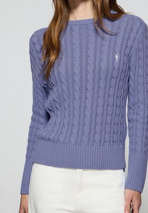 Kobieta ubrana w lawendowy sweter z warkoczowym splotem, z prążkowanymi mankietami i dołem, w połączeniu z białymi spodniami, pokazana od szyi do bioder.