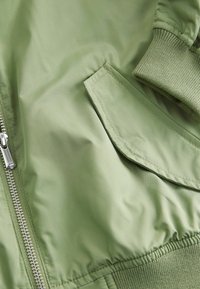 Olivno zelena bomber jakna z gladkim zaključkom, ki ima zadrgo, rebraste manšete in sprednji žep z detajlom lopute.