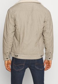 Beige kordjacka med en mjuk krage, knappade ärmslut och sidofickor, visad bakifrån mot en vit bakgrund.