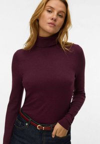 Maglione a collo alto viola realizzato in tessuto morbido, con maniche lunghe e una vestibilità aderente, abbinato a jeans in denim scuri e una cintura rossa.