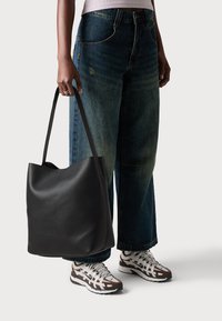 En person som bär vida jeans och vita sneakers håller en stor svart läderaxelväska vid sin sida.