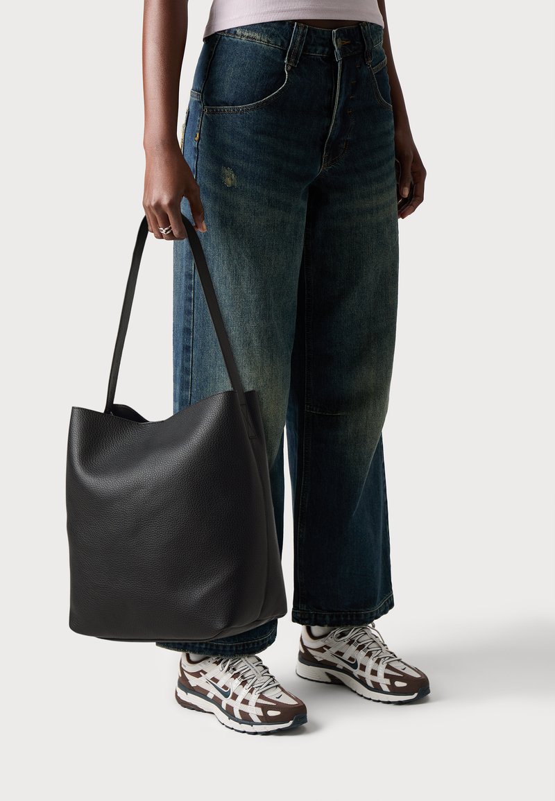 En person som bär vida jeans och vita sneakers håller en stor svart läderaxelväska vid sin sida.