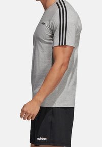 Uomo che indossa una t-shirt grigia Adidas con tre strisce nere sulle maniche e pantaloni corti neri Adidas, in piedi di lato su uno sfondo bianco.