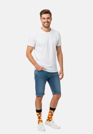 Weißes Baumwoll-T-Shirt, blaue Jeansshorts, weiße Turnschuhe und bunte Socken mit Obstmustern. Stehende Pose mit der Hand in der Tasche.