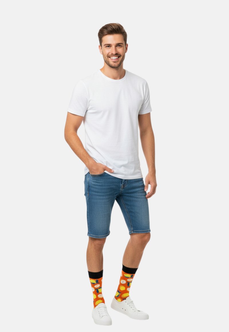 Weißes Baumwoll-T-Shirt, blaue Jeansshorts, weiße Turnschuhe und bunte Socken mit Obstmustern. Stehende Pose mit der Hand in der Tasche.