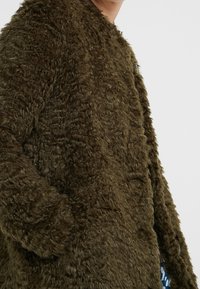Faux-Fur-Mantel in olivgrün, mit einer flauschigen Textur, offenem Design und seitlichen Taschen. Geeignet für den Freizeitlook.
