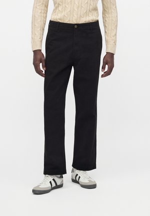 Mand iført sorte bukser med lige ben, hvide og sorte sneakers og en beige kabelstrikket sweater, stående mod en ensfarvet baggrund.