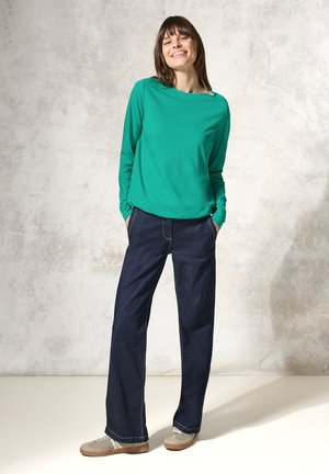 Groene longsluggenshirt met een ontspannen pasvorm, gecombineerd met donkerblauwe wijde jeans. Model draagt lichtgrijze sneakers en staat met handen in de zakken.
