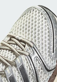 Bílé síťované sportovní boty se stříbrnými akcenty a béžovými tkaničkami, ukazující detailní texturu a přední horní část.