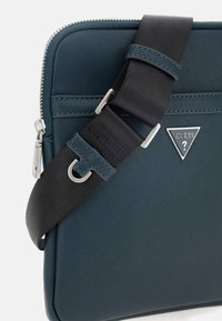 Borsa sintetica verde scuro con chiusura a zip, tracolla nera regolabile e accessori in argento, caratterizzata da un dettaglio logo triangolare.