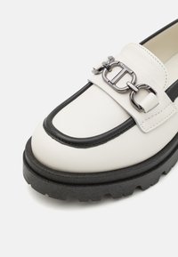 Hvit skinnloafers med svart kant, med en kraftig svart såle og et sølvkjede som pynt på stroppen. Teksturert overflate, moderne design.