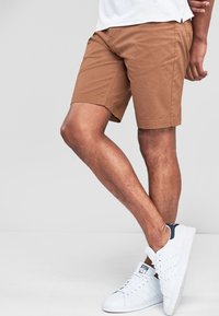 Shorts en coton marron à coupe slim, avec un devant plat, des poches latérales et un design simple. Portés avec des baskets blanches.