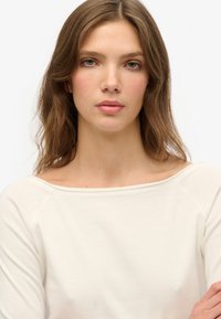 Lys beige top med en bred udskæring og raglanærmer, lavet af blødt stof. Glat tekstur, uden synlige mønstre eller accenter.