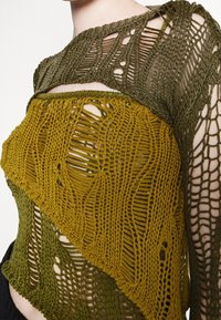 Strikket pullover i olivengrønne og sennepsgule farver, med et tekstureret, åbent strikkemønster og asymmetrisk design.