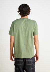 Hombre con cabello oscuro y ondulado, vistiendo una camisa de manga corta verde oliva sencilla y pantalones negros, de pie y de espaldas contra un fondo blanco.