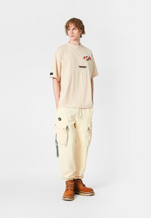 Camiseta oversized beige con detalles bordados, combinada con pantalones cargo de color crema claro que tienen bolsillos con cremallera y botas de color óxido.