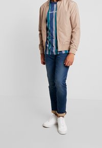 béžová bomber bunda, modro-zelené pruhované tričko, modré džíny a bílé tenisky. Bunda má zipový uzávěr a žebrované náplety.