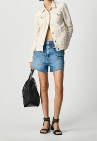 Light beige denim jacket, blue high-waisted shorts, and black studded-strap sandals. Black leather drawstring bag with metal accents.