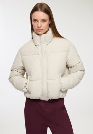 Donna che indossa un giubbotto imbottito crema con colletto alto e mani nelle tasche, abbinato a pantaloni viola scuro su uno sfondo bianco.