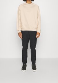 Sudadera beige con el logotipo "HUGO" en relieve, combinada con pantalones oscuros. La sudadera tiene un cuello redondo y puños acanalados. Zapatillas negras completan el conjunto.