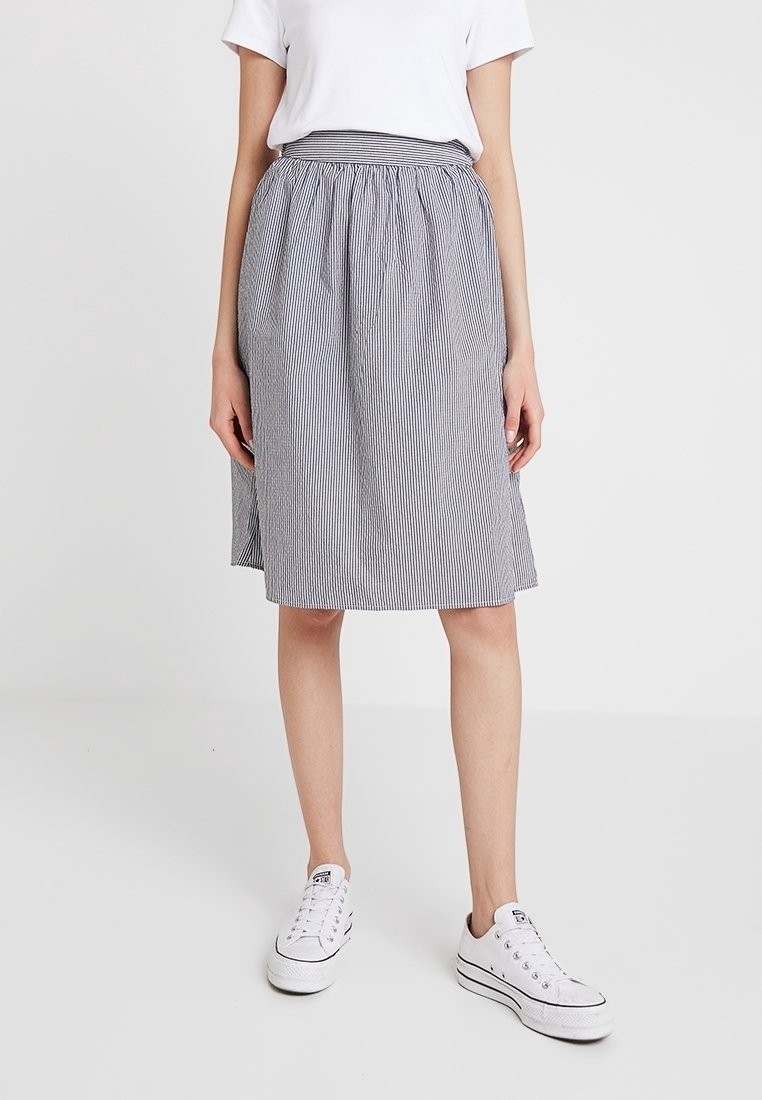 Femme portant une jupe rayée bleu et blanc mi-longue, un haut blanc à manches courtes et des baskets blanches, debout sur un fond uni.