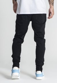 Jeans ajustados negros con una textura suave, con bolsillos traseros. Combinados con zapatillas azul y blancas, sobre un fondo neutro.