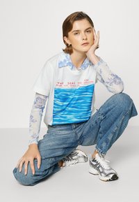 T-shirt branca com gráfico ondulado azul e texto vermelho, sobreposta a uma camisa de manga longa azul com padrão floral, combinada com jeans azuis e ténis prateados.