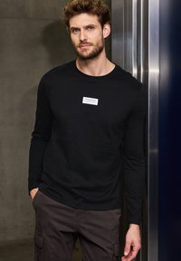 Black long-sleeve t-shirt made of cotton, featuring a rectangular white label with black text on the chest, paired with dark cargo pants.