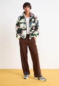 Fuzzy, flerfärgad camouflagjacka med hög krage, kombinerad med lösa bruna byxor med blå sömmar och vita sneakers.