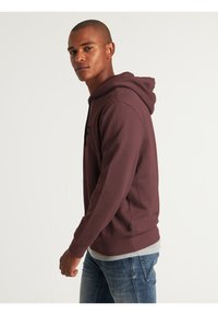 Sudadera burdeos con un acabado texturizado, capucha con cordón y laterales abiertos; llevada sobre una camiseta gris, combinada con jeans azules.