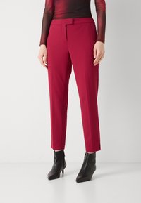 Rode op maat gemaakte broek met een gladde textuur, voorzien van een ceintuur in de taille en een slim fit. Gecombineerd met zwarte enkellaarzen, wat de contrast van de outfit versterkt.
