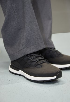 Schwarze Sportschuhe mit strukturiertem Obermaterial, Schnürdesign und weißer Mittelsohle. Getragen mit grauen, halbhohen Hosen und stehend auf einem gesprenkelten Boden.