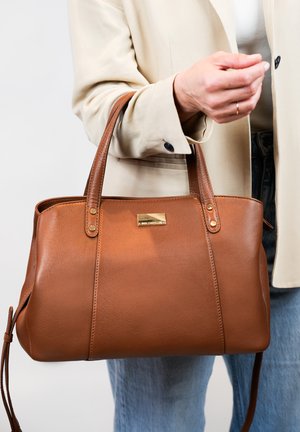 Bruine leren tote bag met accentstiksels en een gouden embleem. Heeft stevige handvatten en een gladde textuur, vastgehouden door een persoon in lichte kleding.
