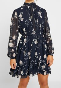 Modellen visar en marinblå blommig klänning med långa ärmar och en volangkant, med hög krage och midjeband. Elegant och feminin stil.