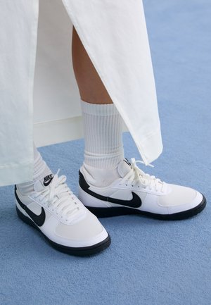 Zapatillas Nike blancas con acentos negros, que presentan un upper de cuero suave, cordones blancos y suelas de goma resistentes, combinadas con calcetines blancos acanalados.